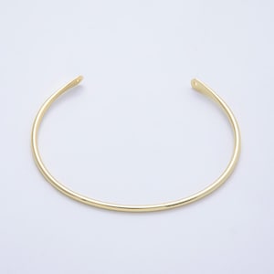 May include: A gold-colored, open-ended cuff bracelet. The bracelet has a smooth, rounded design and tapers slightly at the ends, each with a small hole. The jewelry is set against a plain, light background.
