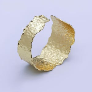 Könnte beinhalten: Ein goldfarbenes Armreif-Armband mit strukturierter, gehammerter Oberfläche. Das offene Design hat eine unregelmäßige, organische Form. Dieses Schmuckstück eignet sich für den Alltag oder besondere Anlässe.