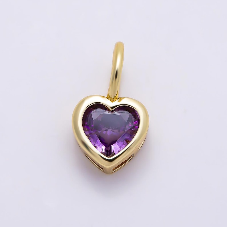 May include: A gold heart-shaped pendant featuring a faceted purple gemstone set in the center. The pendant includes a small gold bail at the top for attachment to a chain, showcasing a polished metallic finish and a deep violet stone.