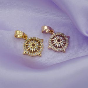 May include: Two gold pendants with a circular design featuring a sunburst pattern with clear rhinestones. The pendant on the left is a bright gold color, while the pendant on the right is a rose gold color.