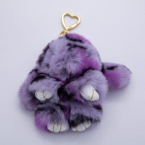 May include: A fluffy purple and black rabbit keychain with a gold-colored heart-shaped clasp. The keychain has white leather-like paws with gold stitching. The rabbit's fur is a mix of purple, black, and white.