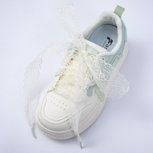 May include: White sneakers with lace-up closure and decorative lace bow. The shoes have a light blue and green accent design. The sole is white and the laces are a cream color. The sneakers have a casual, feminine style.
