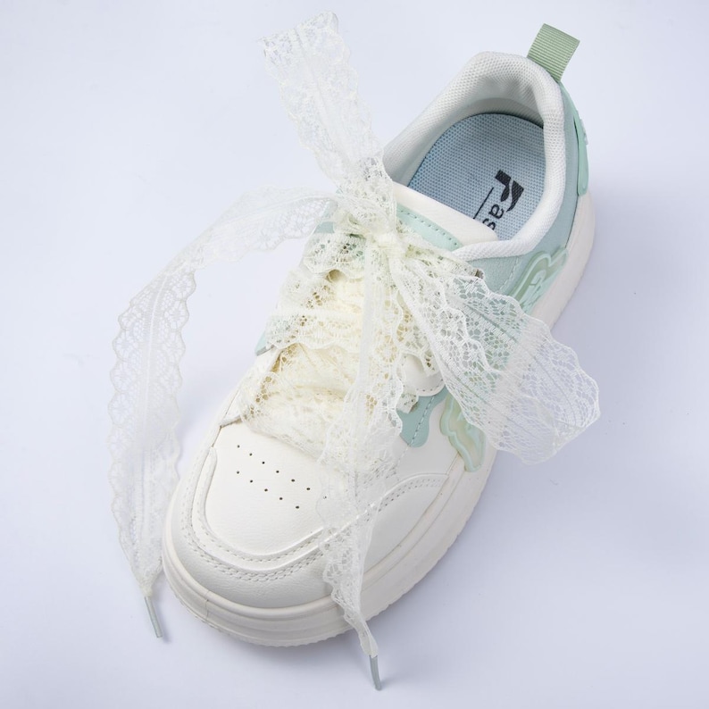 May include: White sneakers with lace-up closure and decorative lace bow. The shoes have a light blue and green accent design. The sole is white and the laces are a cream color. The sneakers have a casual, feminine style.