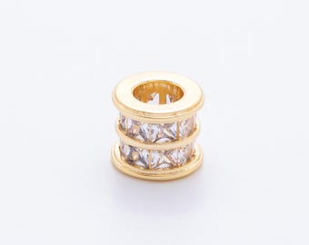 May include: A gold-colored spacer bead with three rows of square, clear crystals. The bead has a cylindrical shape with a hole running through the center. The bead is set against a white background.