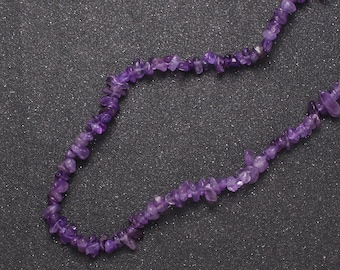 Könnte beinhalten: Eine Halskette aus violetten Amethyst-Edelsteinen, die aus kleinen, unregelmäßig geformten Perlen besteht. Die Halskette liegt auf einem dunkelgrauen Hintergrund.