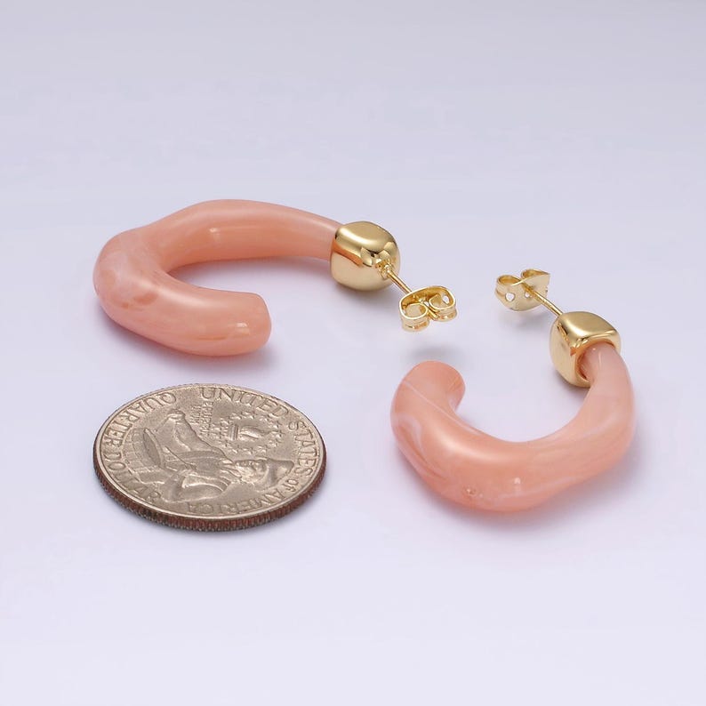 May include: A pair of peach-colored, curved hoop earrings with gold-colored posts and backs. The earrings are next to a U.S. quarter coin. The earrings have a marbled appearance.