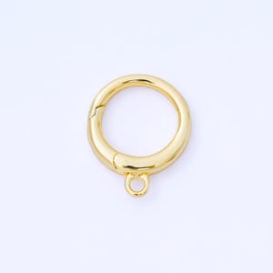 May include: A gold-colored, round clasp with a hinged opening and a small loop at the bottom. The clasp is designed to secure items and is likely used in jewelry making or crafting projects. The metal has a shiny finish.