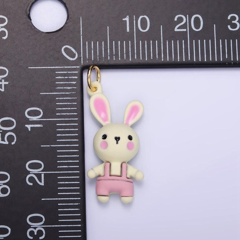 May include: A gold charm featuring a white bunny wearing pink overalls. The bunny has pink ears and a cute face with black eyes and pink cheeks.