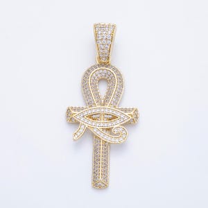 May include: Gold-toned pendant featuring an Ankh cross design, embellished with numerous small, clear gemstones. The pendant includes an eye of Horus symbol and a bail for a chain. The jewelry piece is set against a white background.