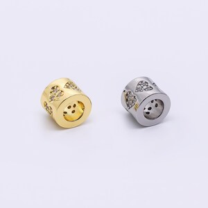 May include: Two cylindrical beads, one gold-toned and one silver-toned, are displayed. Each bead features heart-shaped cutouts filled with small, clear stones. The beads have a hollow center and are likely used for jewelry making or crafting.