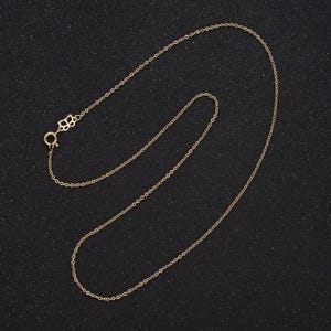 May include: A delicate gold-colored necklace with a small, decorative clasp. The chain is thin and appears to be made of small, connected links. The necklace is displayed on a dark, textured surface, highlighting its simple elegance.