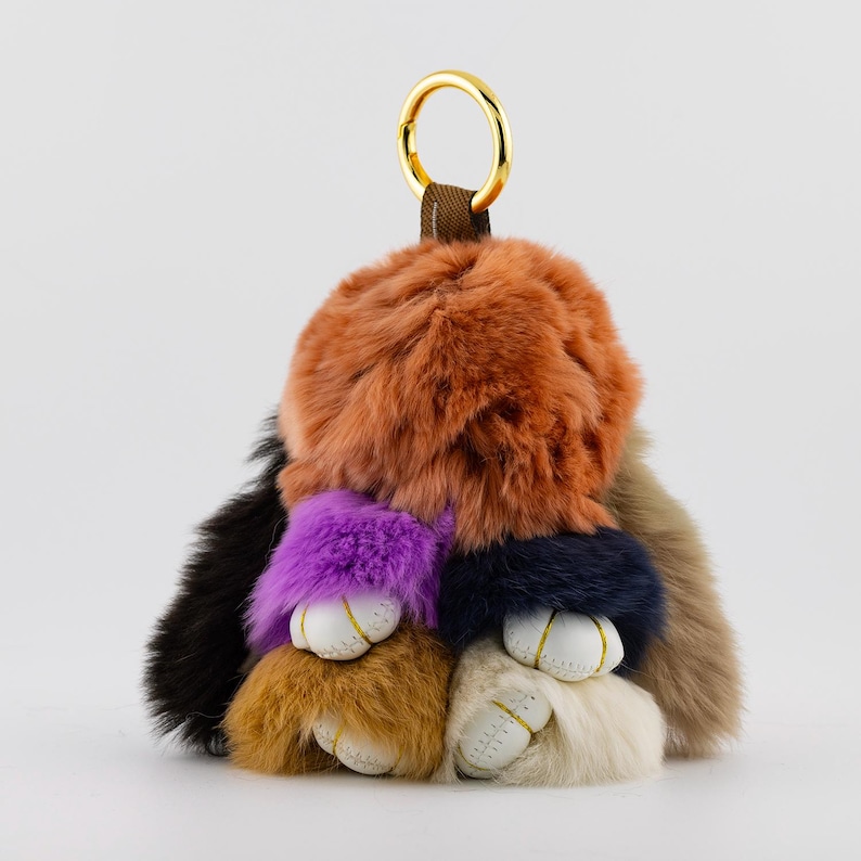 May include: A colorful fur keychain with a gold-tone ring. The keychain features a mix of orange, black, purple, navy, tan, and cream-colored fur. Small white leather accents with gold stitching are visible.