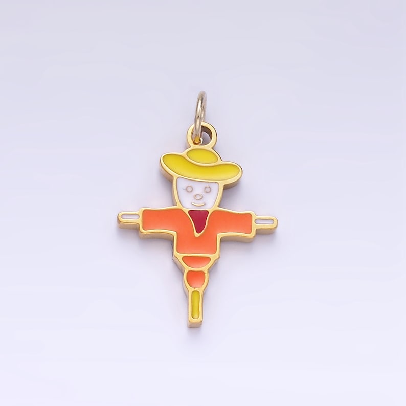 Op de afbeelding: Een gouden hanger in de vorm van een cartoon-vogelverschrikker. De vogelverschrikker draagt ​​een gele hoed, een oranje shirt en een rode sjaal. De hanger heeft een lus aan de bovenkant om aan een ketting of armband te bevestigen.