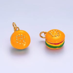 May include: Two gold-tone hamburger charm pendants. One charm is a simple yellow bun with sesame seeds. The other charm is a layered hamburger with yellow bun, red tomato, green lettuce, and yellow cheese.