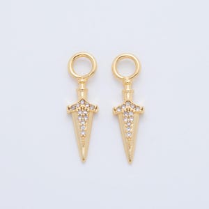 May include: Two gold-colored dagger-shaped charms with a loop at the top for hanging. The daggers are embellished with small, clear crystals along the blade and at the hilt, creating a sparkling effect. The background is a plain, light color.