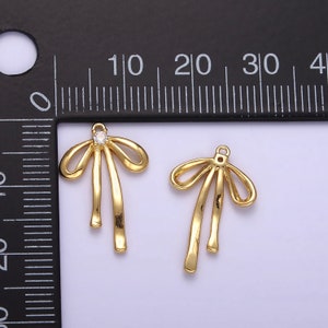 K&ouml;nnte beinhalten: Zwei goldfarbene Schleifen-Anh&auml;nger mit einem klaren Strassstein in der Mitte der Schleife. Die Anh&auml;nger sind etwa 2 cm lang.
