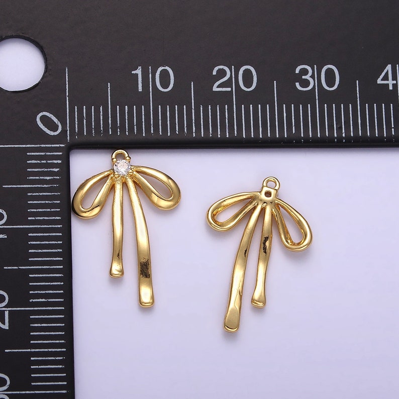 K&ouml;nnte beinhalten: Zwei goldfarbene Schleifen-Anh&auml;nger mit einem klaren Strassstein in der Mitte der Schleife. Die Anh&auml;nger sind etwa 2 cm lang.
