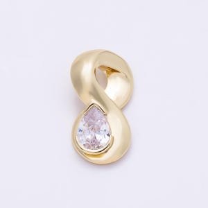 Pode incluir: Um pingente de ouro em forma de símbolo de infinito estilizado com um acabamento liso e polido. Uma única pedra preciosa transparente em forma de pera e facetada está engastada na curva inferior da armação de ouro, criando um contraste.
