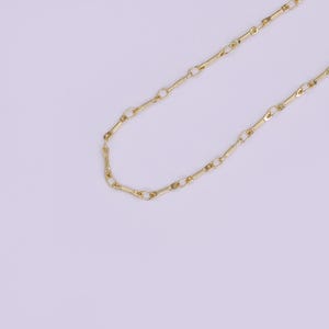 May include: A delicate gold-colored chain necklace. The chain is composed of elongated, oval-shaped links connected by smaller, circular links. The necklace is displayed against a soft, light purple background.