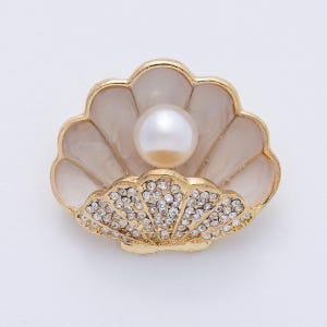 May include: A gold-toned seashell brooch with a large, round, white pearl at the center. The shell has a cream-colored enamel finish and is encrusted with small, sparkling crystals.