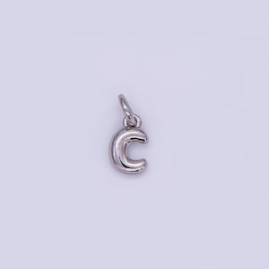 May include: A silver-colored charm in the shape of the letter "C". The charm has a rounded, three-dimensional design with a small loop at the top for attaching to a necklace or bracelet. The background is a light, neutral color.