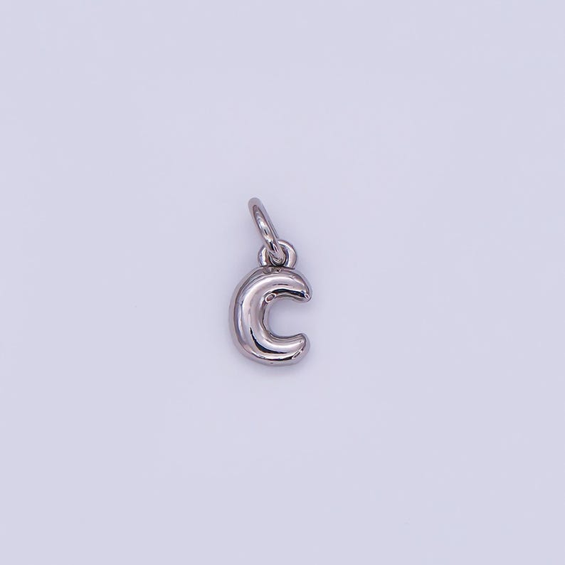 May include: A silver-colored charm in the shape of the letter "C". The charm has a rounded, three-dimensional design with a small loop at the top for attaching to a necklace or bracelet. The background is a light, neutral color.