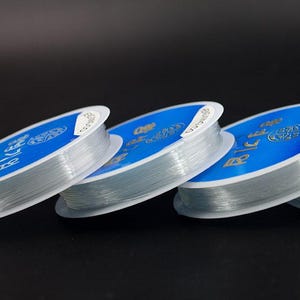 May include: Four spools of clear elastic string wound around blue and white labels. The labels have text in English and Japanese. The spools are stacked on top of each other.