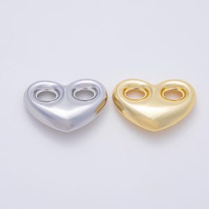 May include: Two heart-shaped buttons with a smooth, metallic finish. One button is silver-toned and the other is gold-toned. Each heart features two circular holes positioned in the upper center for attachment, with a rounded, puffed surface design.