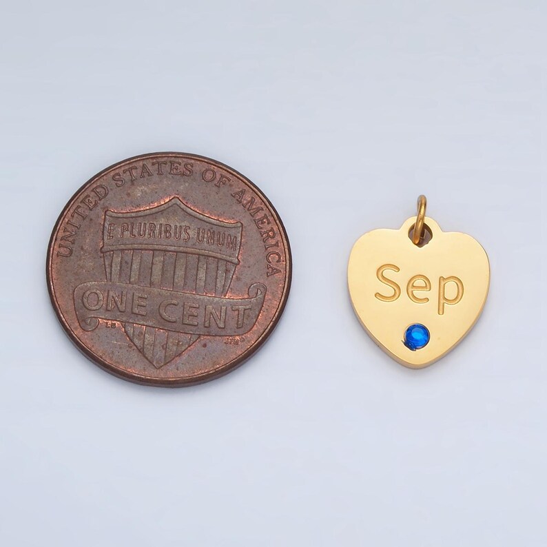 May include: Gold heart-shaped charm with the word "Sep" engraved on it. The charm has a small blue gemstone in the center. The charm is shown next to a US penny for size comparison.