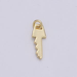 May include: A gold-colored key charm with a simple design. The key has a rounded top and a serrated edge.