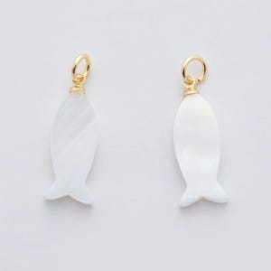 May include: Two white fish-shaped charms with a pearlescent sheen. Each charm has a gold-colored loop at the top for hanging. The charms are designed with a tail fin and a rounded body.