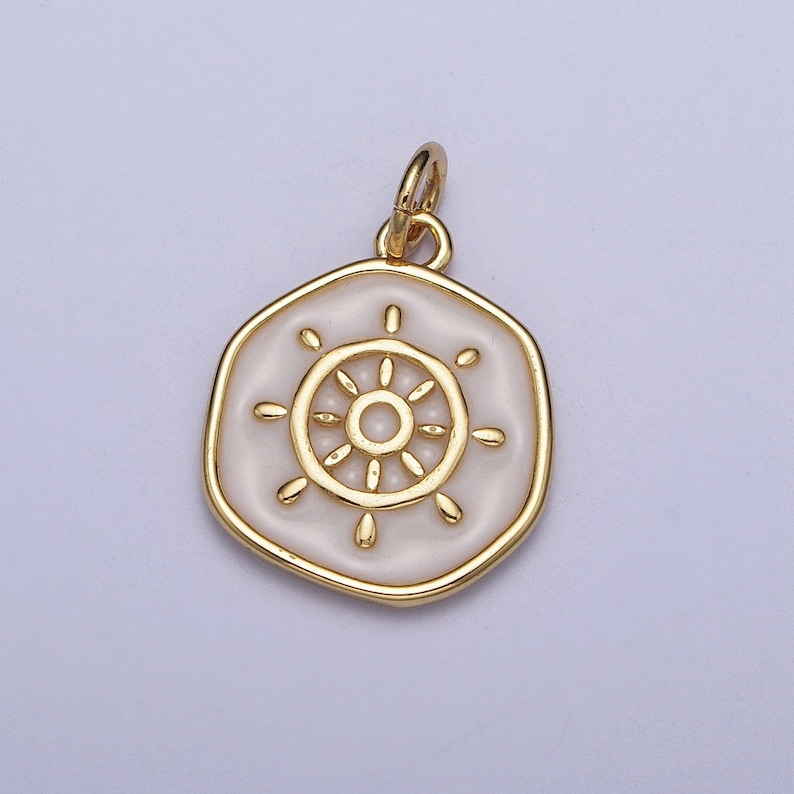K&ouml;nnte beinhalten: Goldfarbener Anh&auml;nger mit wei&szlig;em Emaille-Mittelteil, das ein Steuerraddesign zeigt. Der Anh&auml;nger hat eine sechseckige Form mit einem goldenen Rand und einer kleinen Schlaufe f&uuml;r eine Kette. Das Raddesign ist ebenfalls goldfarben.