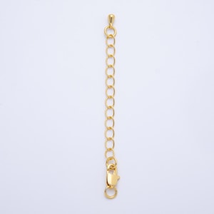 May include: A gold-colored necklace extender with a lobster clasp and a small, round end piece. The extender is made of a series of oval-shaped links. The background is plain white.