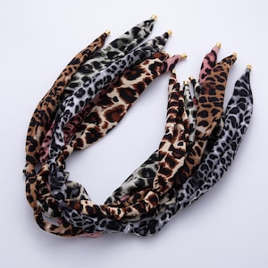 May include: A collection of fabric headbands with various leopard print patterns. The headbands feature a twisted design and gold-colored metal tips. The prints include brown, black, gray, and pink leopard spots on different colored backgrounds.
