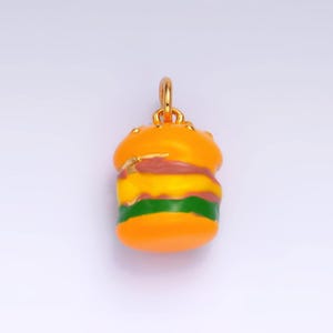 May include: A gold charm shaped like a hamburger with yellow, green, and brown layers. The charm is a fun and quirky accessory for a necklace or bracelet.