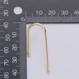 May include: A gold-colored, U-shaped jewelry finding with a curved top and straight sides. The finding has small loops at the bottom of each straight side. A ruler is in the background.
