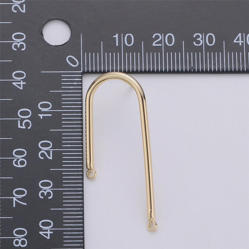 May include: A gold-colored, U-shaped jewelry finding with a curved top and straight sides. The finding has small loops at the bottom of each straight side. A ruler is in the background.