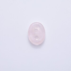 May include: A pale pink, oval-shaped button with two holes. The button is made of a translucent material, possibly a gemstone or glass, and has a smooth, polished surface. The button is set against a white background.
