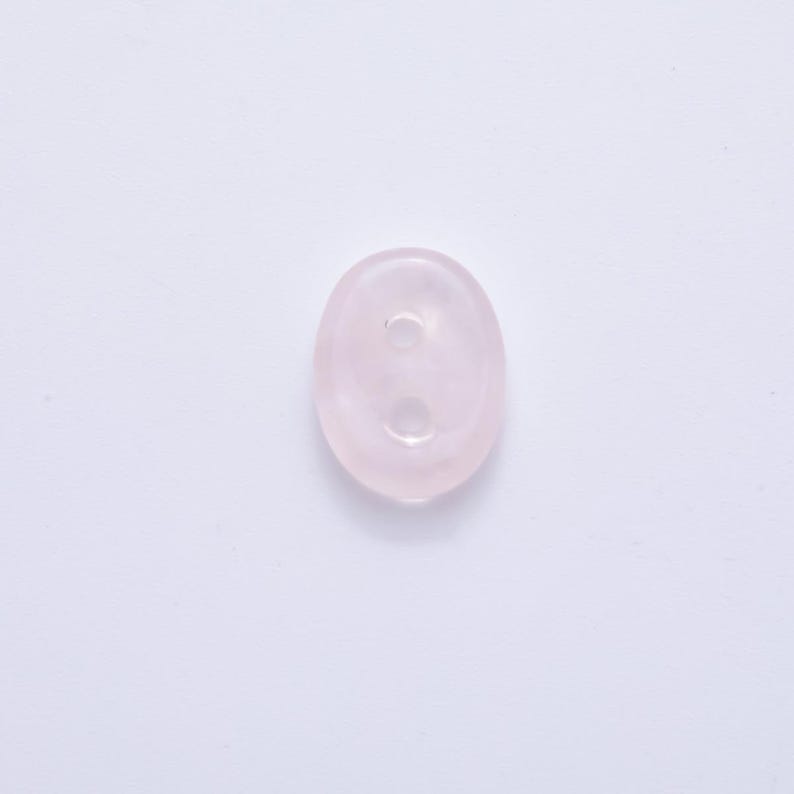 May include: A pale pink, oval-shaped button with two holes. The button is made of a translucent material, possibly a gemstone or glass, and has a smooth, polished surface. The button is set against a white background.