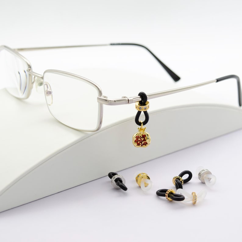 Puede incluir: Un par de gafas con montura plateada, una correa de goma negra y un dije de oro en forma de granada con piedras de imitaci&oacute;n rojas y transparentes. El dije est&aacute; unido a la correa con un anillo de plata. Tambi&eacute;n hay varias correas de goma negra con anillos de plata y piedras de imitaci&oacute;n transparentes sobre una superficie blanca.