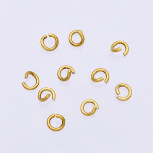 May include: A collection of small, open jump rings in a bright gold color. These jewelry-making components are scattered against a white background. The rings are circular with a small opening for attaching other jewelry pieces.