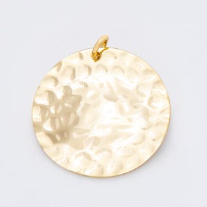 Puede incluir: Un colgante dorado con textura martillada de forma circular. El colgante tiene un pequeño lazo en la parte superior para sujetarlo a una cadena o cordón. La superficie presenta un patrón irregular y con hoyuelos.