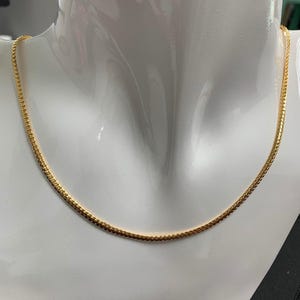 Puede incluir: Un collar de cadena de oro en un maniquí blanco. La cadena es fina y delicada.