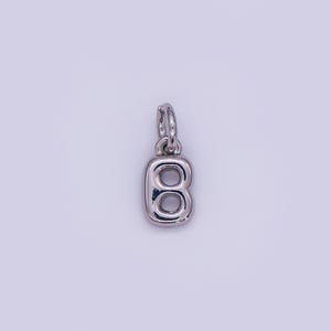May include: A silver-colored charm in the shape of the letter "B". The charm has a loop at the top for attaching to a necklace or bracelet. The letter is stylized with rounded edges and a hollow center.