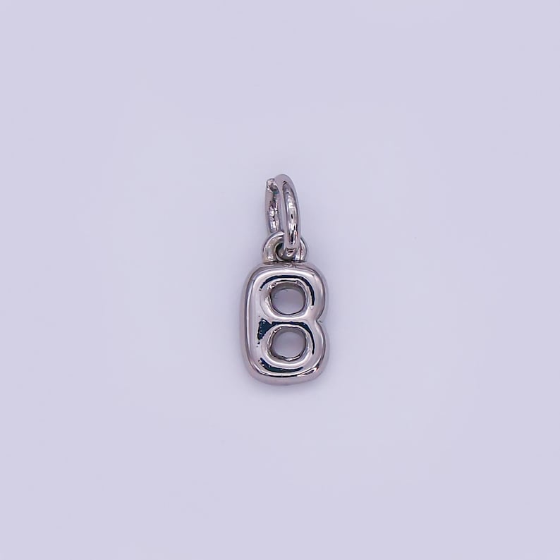 May include: A silver-colored charm in the shape of the letter "B". The charm has a loop at the top for attaching to a necklace or bracelet. The letter is stylized with rounded edges and a hollow center.