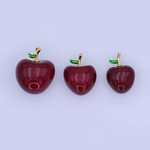 May include: Three glossy, cherry-shaped ornaments in varying sizes. Each cherry is a deep red with a gold stem and a small green leaf. The ornaments are arranged on a plain, light background.