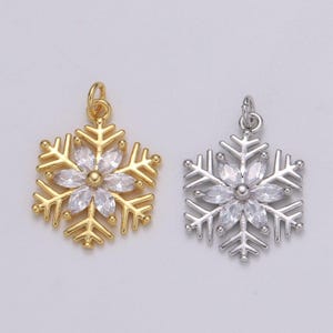 May include: Two snowflake pendants, one gold-toned and one silver-toned. Each pendant features a central flower design with clear, faceted stones. The snowflake design has detailed, pointed edges and a small loop for hanging.