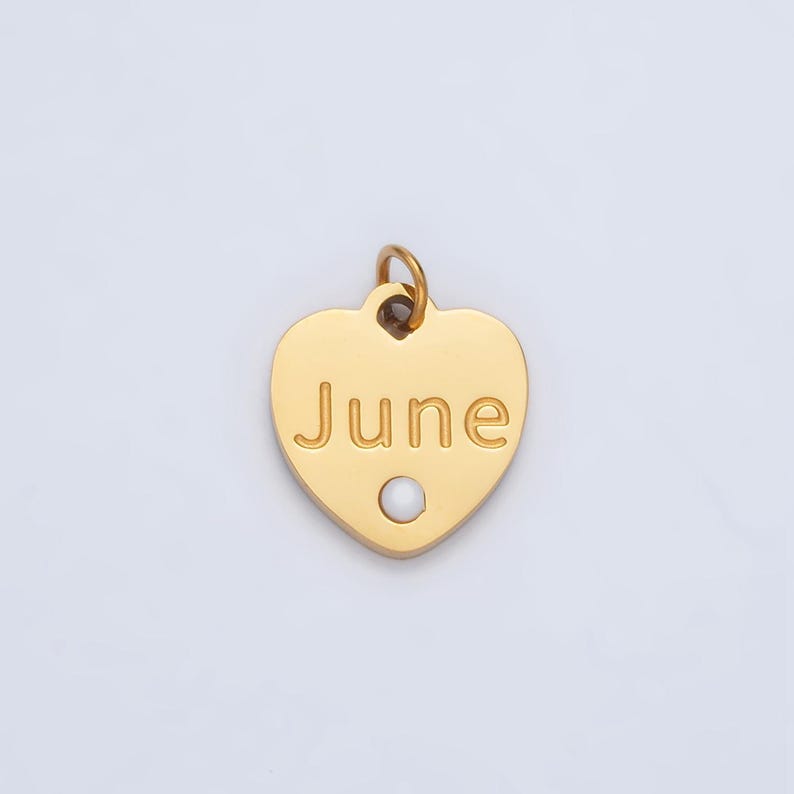 May include: Gold heart-shaped charm with the word "June" engraved on it. A small white pearl is centered on the charm.
