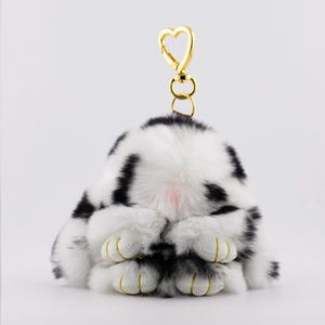 May include: A plush bunny keychain in black and white spotted fur. The bunny is in a seated position with its paws visible. The keychain has a gold-colored heart-shaped clasp.