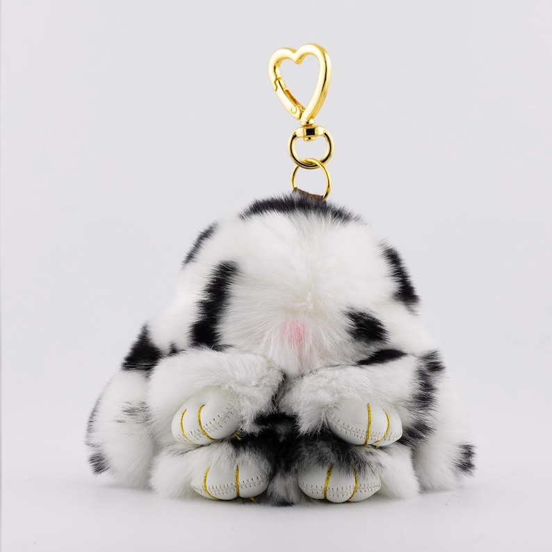 May include: A plush bunny keychain in black and white spotted fur. The bunny is in a seated position with its paws visible. The keychain has a gold-colored heart-shaped clasp.